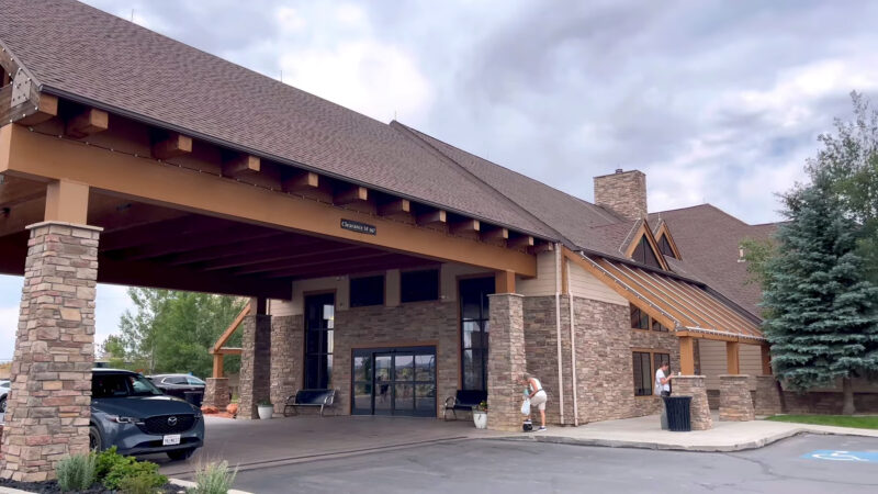 Exterior view of Bryce Canyon Resort with a covered entrance supported by stone columns, featuring a car parked in front. A person is seen picking up items near the entrance, and there are shrubs and trees in the surroundings under a cloudy sky