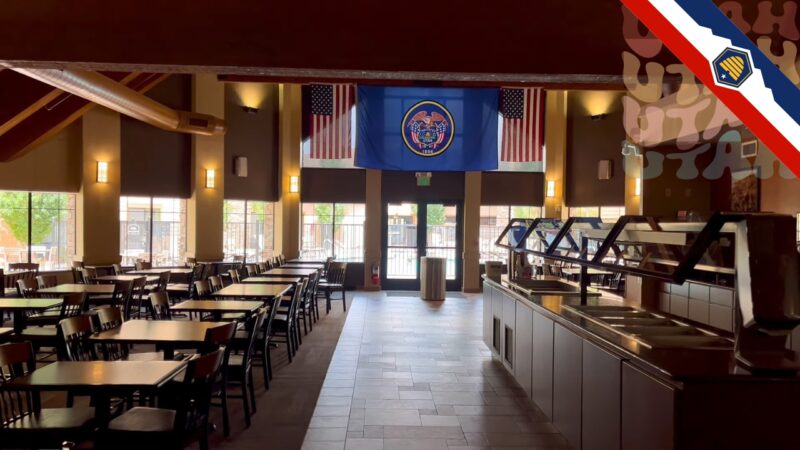 Interior view of a hotel dining area with rows of tables and chairs. The Utah state flag and American flags hang from the ceiling. The entrance is visible in the background, with large windows allowing natural light to fill the space