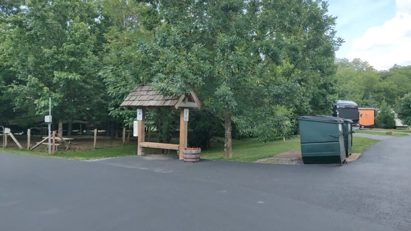 image of Up the Creek RV Park in Pigeon Forge, showcasing a wooden welcome sign under a canopy of trees. The park area features picnic tables and garbage bins, with RVs visible in the background