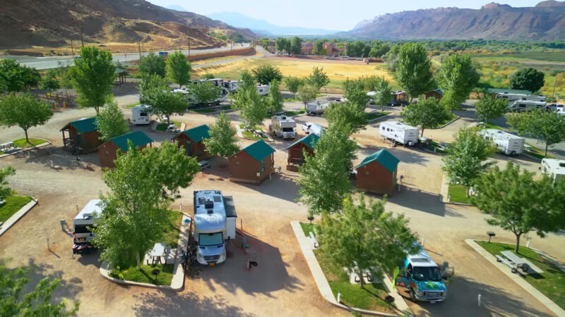 An aerial view of Sun Outdoors Arches Gateway campground in Moab, Utah. The image features several wooden cabins with green roofs, parked RVs, and numerous trees in a well-maintained landscape